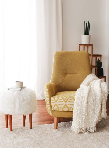 A yellow upholstered armchair with a geometric pattern, positioned in a room with a white curtain, a wooden side table, and a white rug. A white throw blanket is draped over the armchair, and there is a potted plant and a decorative object on the side table.