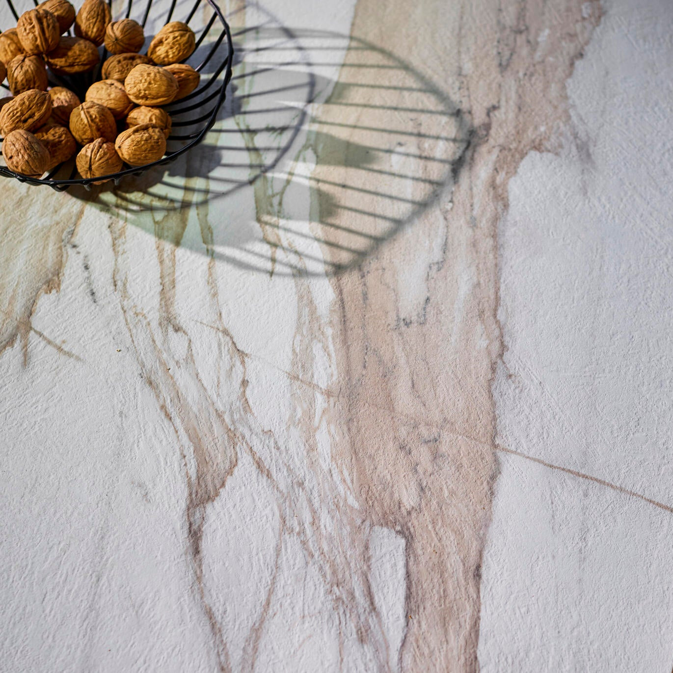 A close-up of a white marble-look tabletop with soft beige and gray veining, styled with a black wire bowl of walnuts and a yellow ceramic cup in the foreground.