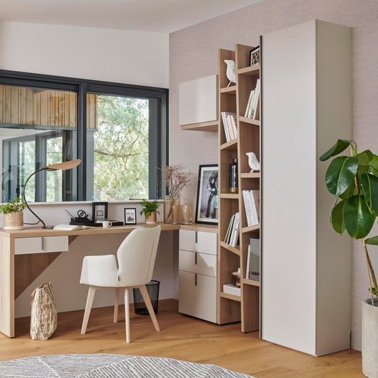 A modern home office setup featuring a light wood desk with a white chair, a tall white storage cabinet, and a slatted wooden bookshelf filled with books and decor, set against a light textured wall with a large window.