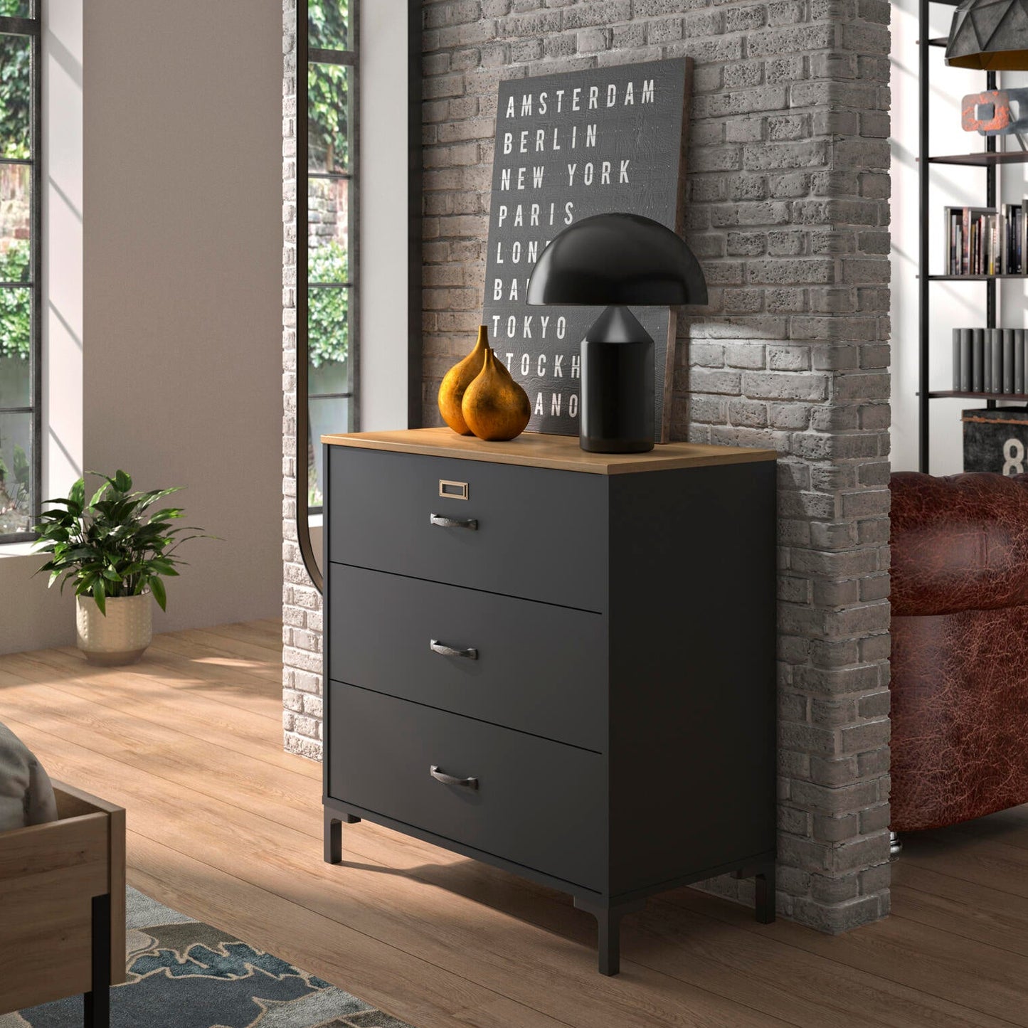 A dark gray three-drawer dresser with a light wood top in a modern bedroom setting, styled with a black table lamp, decorative gourds, and a framed print against a gray brick column.