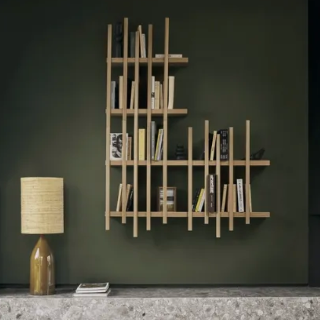A modern wall-mounted wooden bookshelf with an asymmetrical, staggered grid design, styled with books and small decor items against a dark olive green wall.