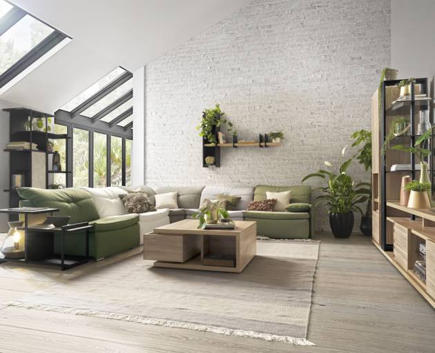 A modern living room featuring a green sectional sofa, a light wood coffee table, a large beige rug, and a white brick accent wall with built-in shelving and plants.