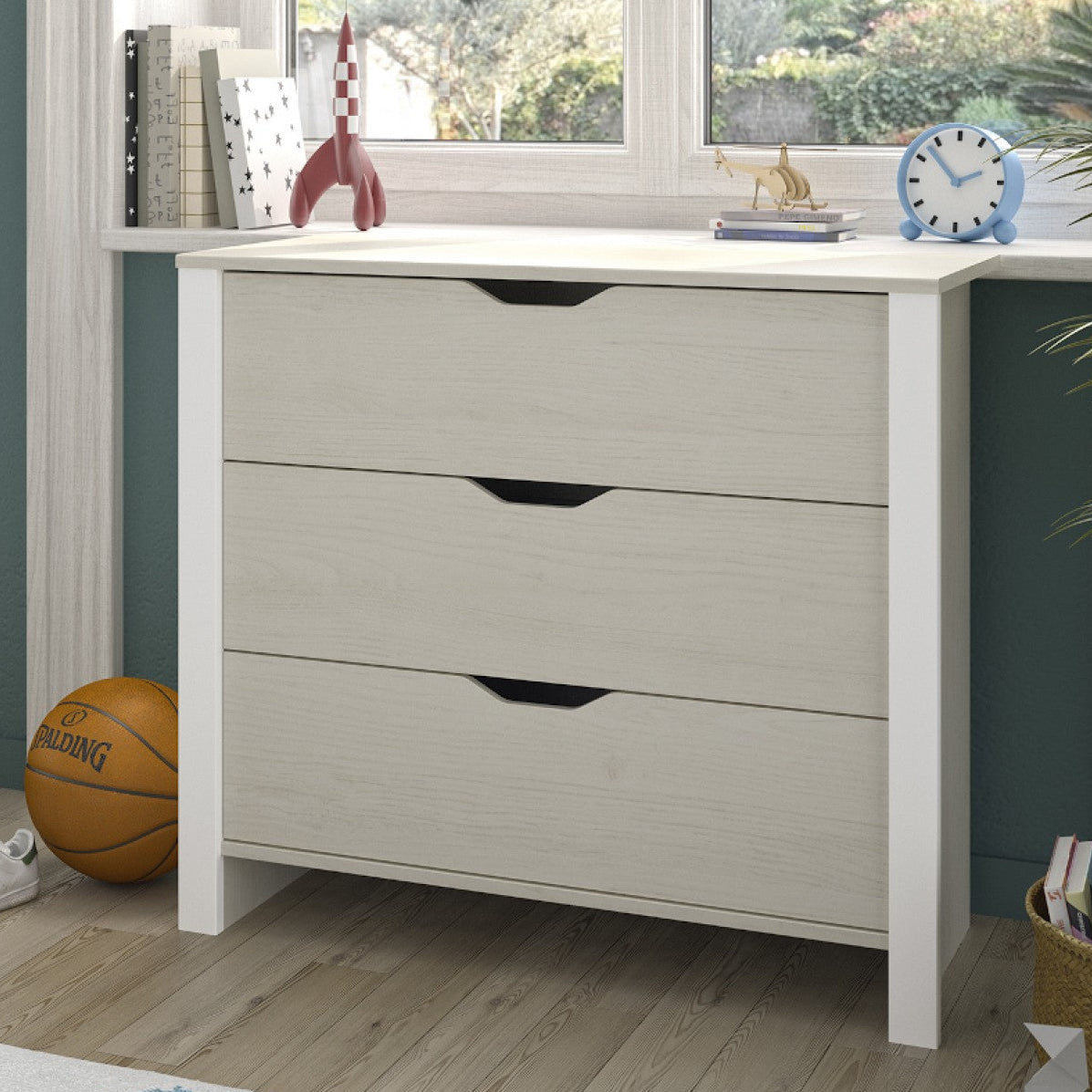 A three-drawer dresser with a light gray wood-grain finish and black drawer pulls, shown in a styled room setting with a window, books, a basketball, and a plant.
